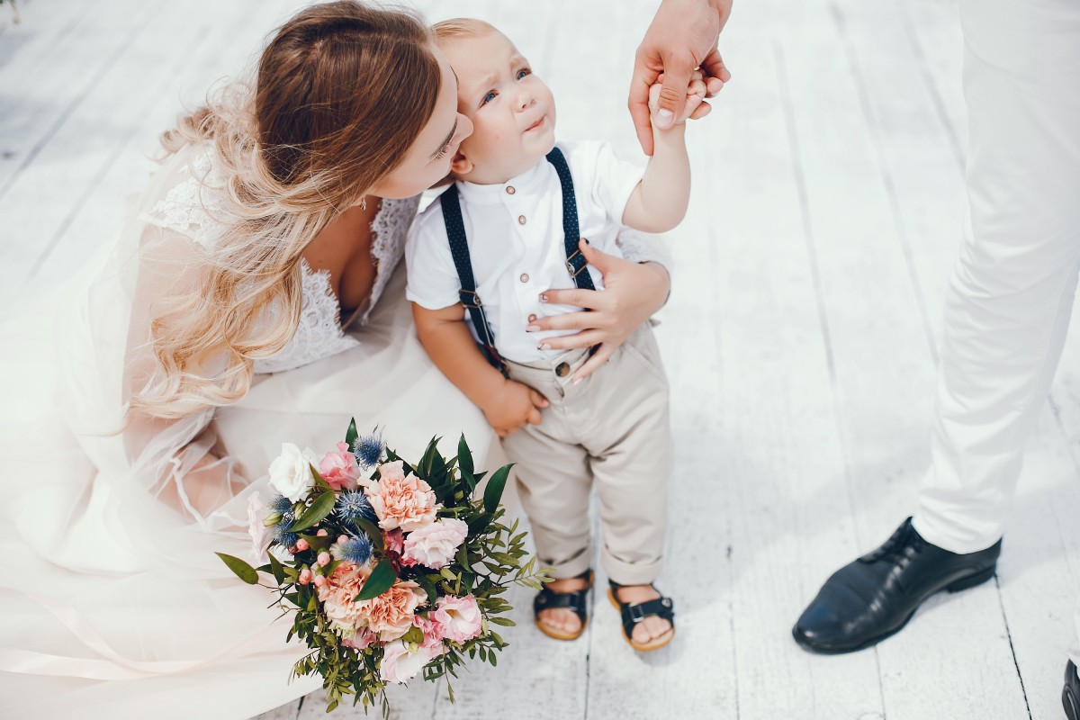 Une mère en robe de cérémonie embrasse tendrement son petit garçon habillé avec élégance, tandis que le père lui tient la main. L’enfant porte un pantalon clair avec bretelles et sandales noires. Un bouquet de fleurs pastel repose au sol sur un parquet blanc.